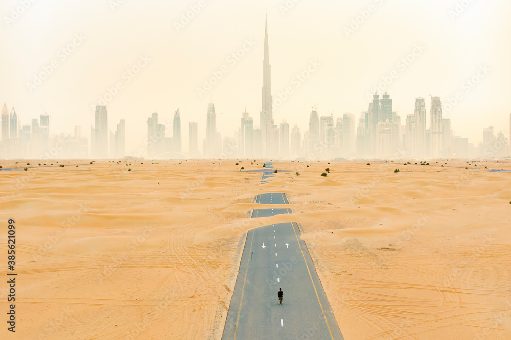 View from above, stunning aerial view of an unidentified person walking on a deserted road covered by sand dunes with the Dubai Skyline in the background. Dubai, United Arab Emirates.