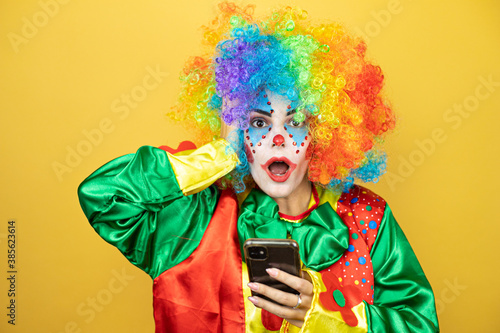 Clown standing over yellow insolated yellow background chatting with her phone and surprised