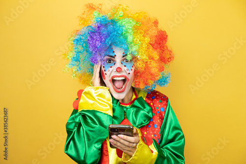 Clown standing over yellow insolated yellow background chatting with her phone and surprised