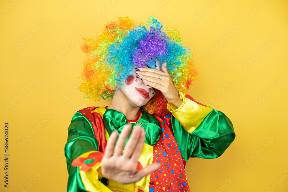 Clown standing over yellow insolated yellow background covering eyes with hands and doing stop gesture with sad and fear expression. Embarrassed and negative concept. Clown standing over yellow insolated yellow background covering eyes with hands and doing stop gesture with sad and fear expression. Embarrassed and negative concept.