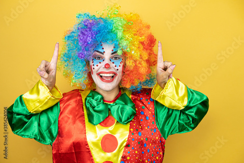 Clown standing over yellow insolated yellow background smiling, looking at the camera and pointing up with fingers and raised arms
