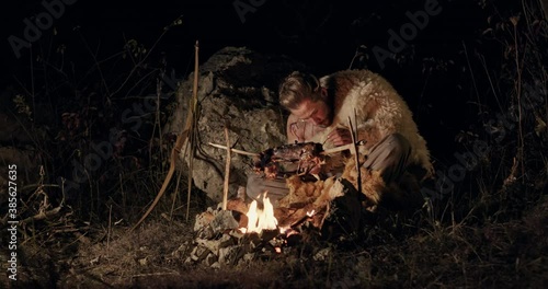 Hungry barbarian eating meat near campfire. Hungry man eating fried meat greedily while resting near bonfire at dark night in nature photo