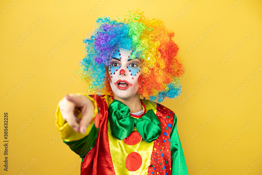 Clown standing over yellow insolated yellow background laughing at you, pointing finger to the camera with hand over body, shame expression Clown standing over yellow insolated yellow background laughing at you, pointing finger to the camera with hand over body, shame expression