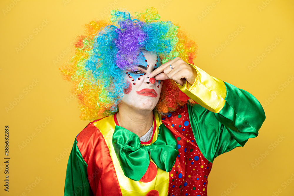 Clown standing over yellow insolated yellow background smelling something stinky and disgusting, intolerable smell, holding breath with fingers on nose Clown standing over yellow insolated yellow background smelling something stinky and disgusting, intolerable smell, holding breath with fingers on nose