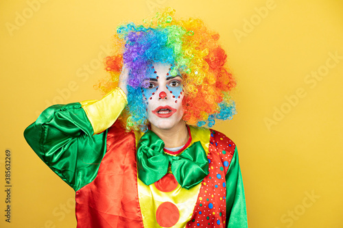 Clown standing over yellow insolated yellow background putting one hand on her head smiling like she had forgotten something