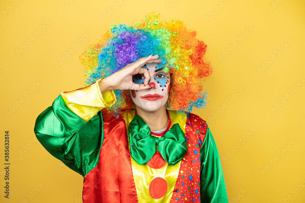 Clown standing over yellow insolated yellow background doing ok gesture shocked with smiling face, eye looking through fingers Clown standing over yellow insolated yellow background doing ok gesture shocked with smiling face, eye looking through fingers