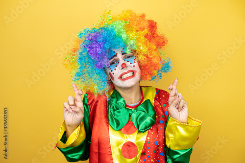 Clown standing over yellow insolated yellow background gesturing finger crossed smiling with hope and looking side