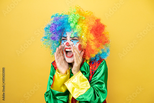 Clown standing over yellow insolated yellow background shouting and screaming loud to side with hands on mouth