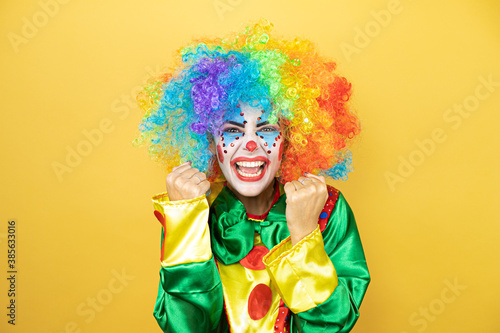 Clown standing over yellow insolated yellow background very happy and excited making winner gesture with raised arms  smiling and screaming for success.