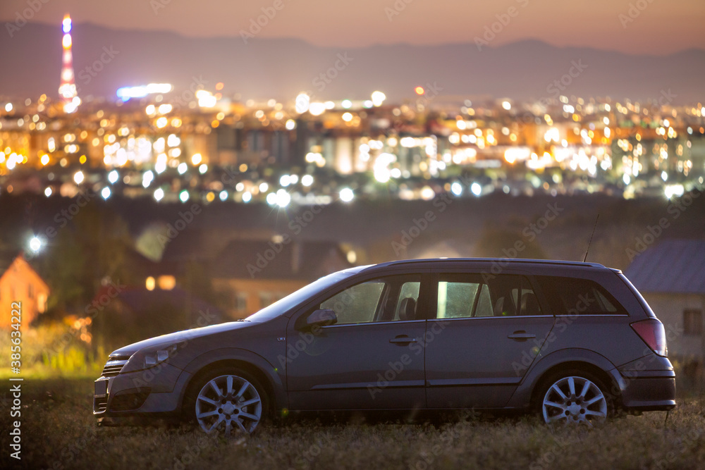 Gray car parked at night in green meadows on background of lights of distant city buildings and dark mountain ridge under bright sky at sunset. Transportation, traveling, vehicles design concept.