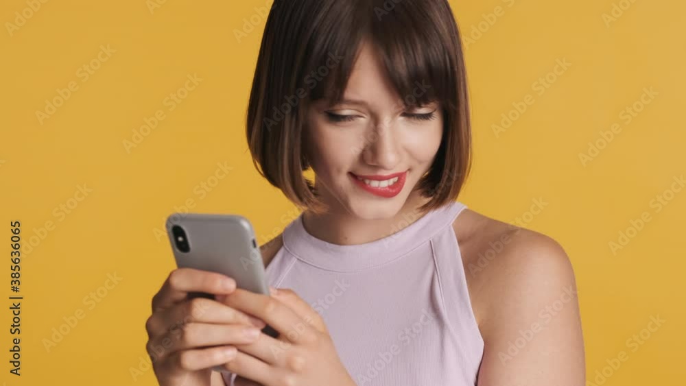 Charming brunette girl with bob hair sensually biting lips chatting with boyfriend on smartphone isolated on yellow background Charming brunette girl with bob hair sensually biting lips chatting with boyfriend on smartphone isolated on yellow background