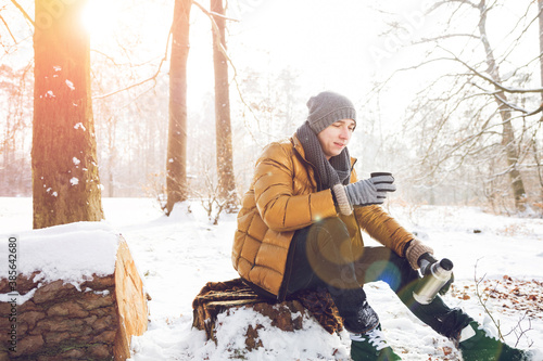 Mann mit einer Thermosflasche heissen Tee  sitzt auf einem Baumstamm im verschneiten  Winter Wald. photo