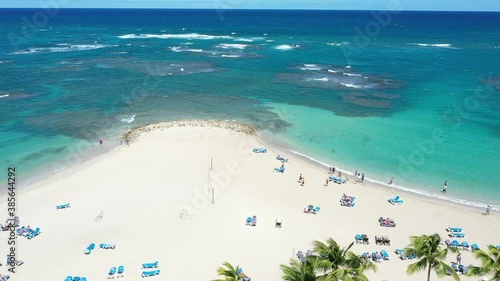 This is a beach with clear blue water and white sand. A drone pan from right to left overlooking palm trees and tourists sunbathing. A small mountain called La Isabela in Puerto Plata, Dom. Rep. photo