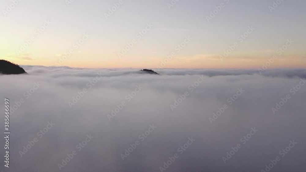 Drone flies over a layer of clouds where the top of a mountain protrudes during a sunrise Drone flies over a layer of clouds where the top of a mountain protrudes during a sunrise