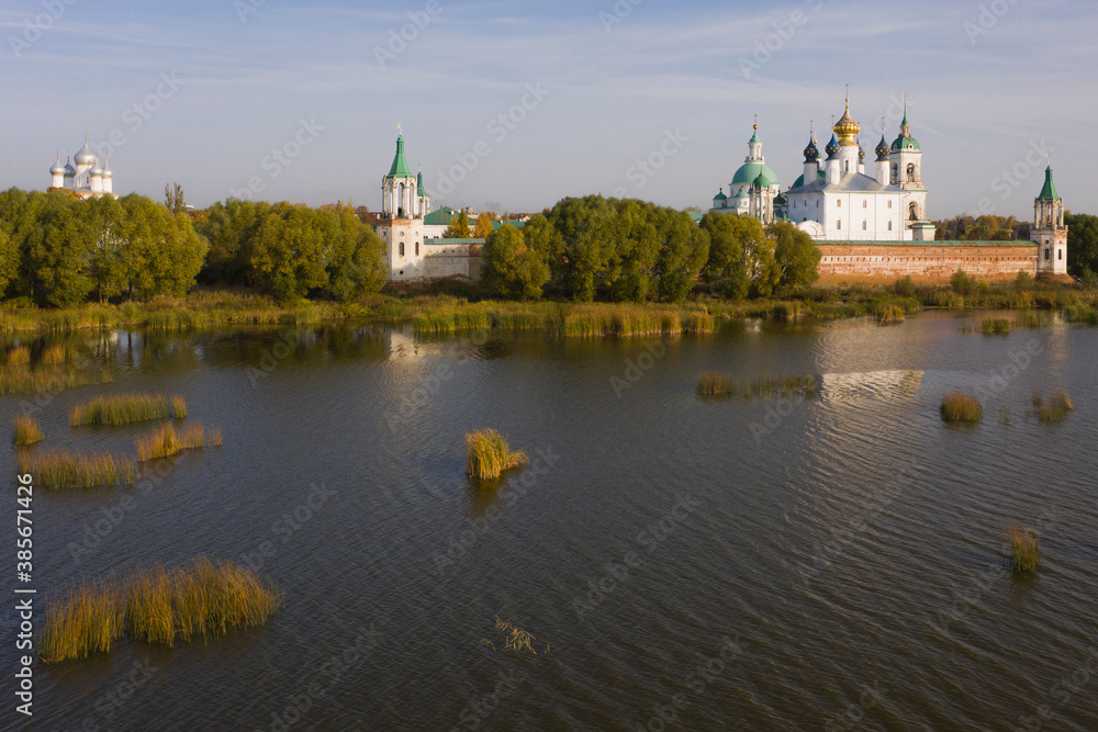 Aerial view of a Russian Orthodox monastery on a lakeside surrounded by grass and trees in autumn colours. Light white clouds on blue sky. No people.