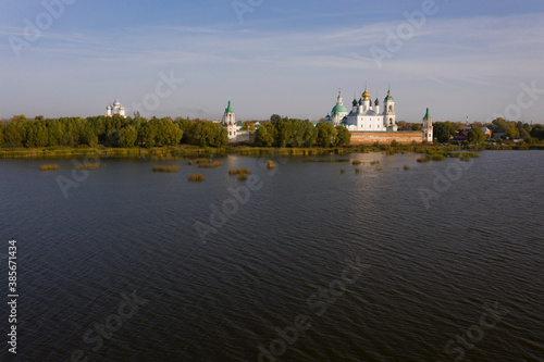 Aerial view of a Russian Orthodox monastery on a lakeside surrounded by grass and trees in autumn colours. Light white clouds on blue sky. No people.