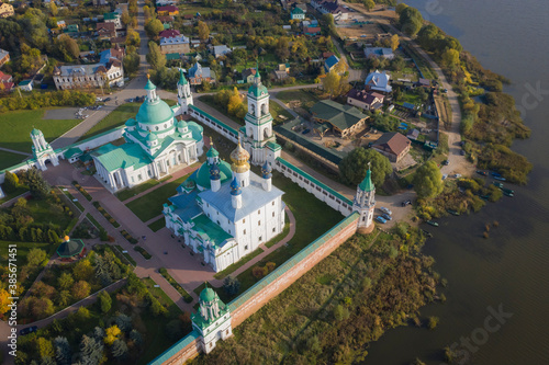 Aerial view of a Russian Orthodox monastery on a lakeside surrounded by grass and trees in autumn colours. Outskirts of a small town. Light white clouds on blue sky.