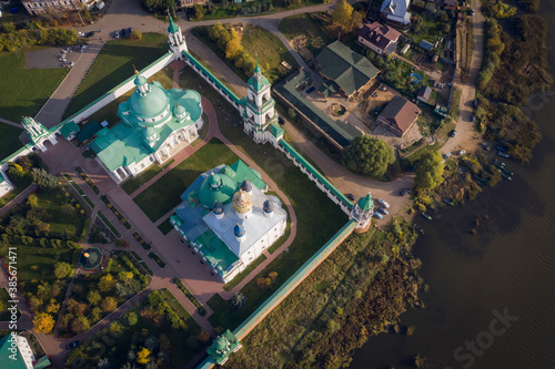 Aerial top-down view of a Russian Orthodox monastery on a lakeside surrounded by grass and trees in autumn colours. Outskirts of a small town. Light white clouds on blue sky.