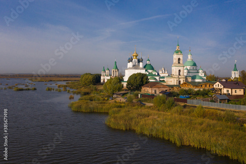 Aerial view of a Russian Orthodox monastery on a lakeside surrounded by grass and trees in autumn colours. Light white clouds on blue sky. No people.