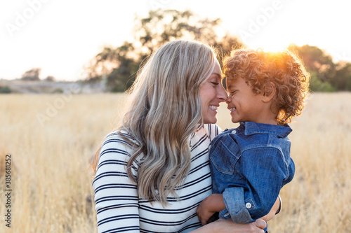 Beautiful happy Mother holding her adorable diverse son in the outdoor sunlight. Sun rays shining through as the mom shows love and affection to her son photo