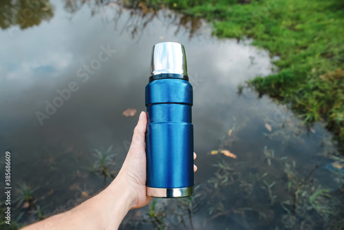 Man holding a hiking flask, lake background. Vacuum metal thermos