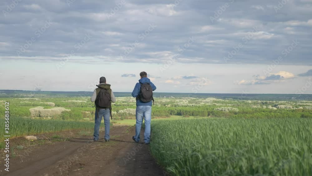 teamwork travel. two man hikers with backpacks walk along a lifestyle trail next to a field with green grass in nature. concept adventure travel hiking healthy. two tourists walk walking talking teamwork travel. two man hikers with backpacks walk along a lifestyle trail next to a field with green grass in nature. concept adventure travel hiking healthy. two tourists walk walking talking