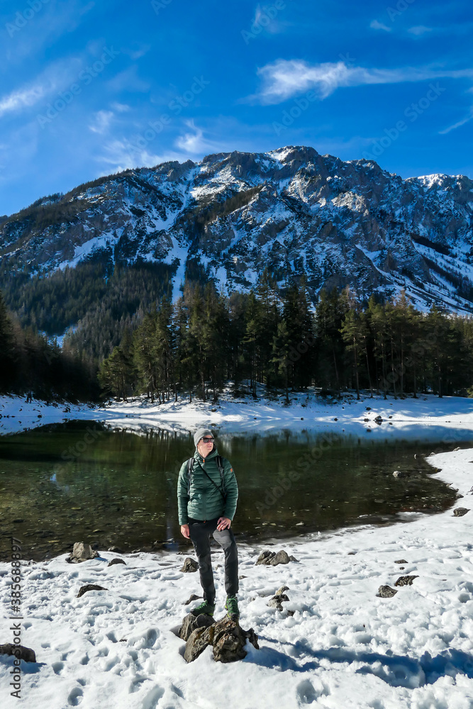 A man standing at the stone next to the shore of Green Lake in Austrian Alps. Mountains and ground are covered with snow. Winter hiking. The man is enjoying a frosty winter day. Winter wonderland