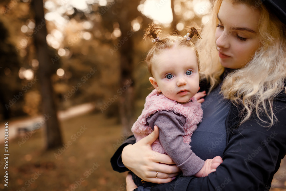Fototapeta premium Young mother is holding her 6 months old baby girl in her arms in the autumn park.