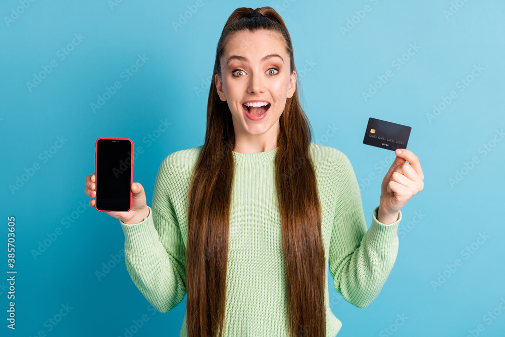 Photo portrait of excited girl with open mouth holding phone with blank space bank card wearing green pullover isolated on pastel blue colored background Photo portrait of excited girl with open mouth holding phone with blank space bank card wearing green pullover isolated on pastel blue colored background