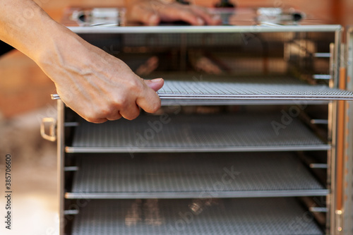 Human hands, taking out empty metal pan from dehydrating machine. Equipment for producing dry meat or vegetarian jerkies. Many mesh shelves inside. Soft selective focus, copy space.