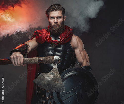 Brutal and armoured roman warrior with red cloak and outstretched arm which holding axe posing in dark room with smoke and spotlight.