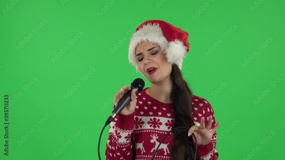 Portrait of sweety girl in Santa Claus hat is singing into a microphone and moving to the beat of music. Green screen Portrait of sweety girl in Santa Claus hat is singing into a microphone and moving to the beat of music. Green screen