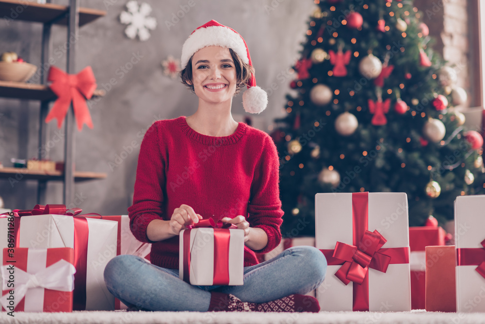 Photo of lovely girl sit carpet receive giftbox toothy smile look camera wear santa cap red pullover decorated living room indoors