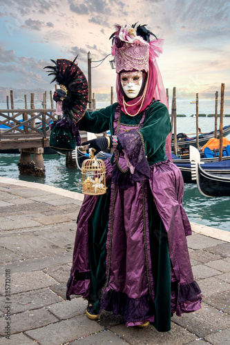 Woman in mask wearing ornate colorful carnival costume on San Marco lagoon background at sunset. Carnival in Venice, Italy
