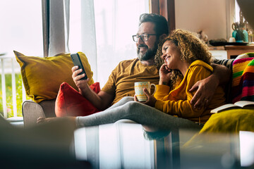 Adult caucasian couple at home enjoy phone call conference with friends - people and technology activity - happy man and woman have breakfast together on the couch
