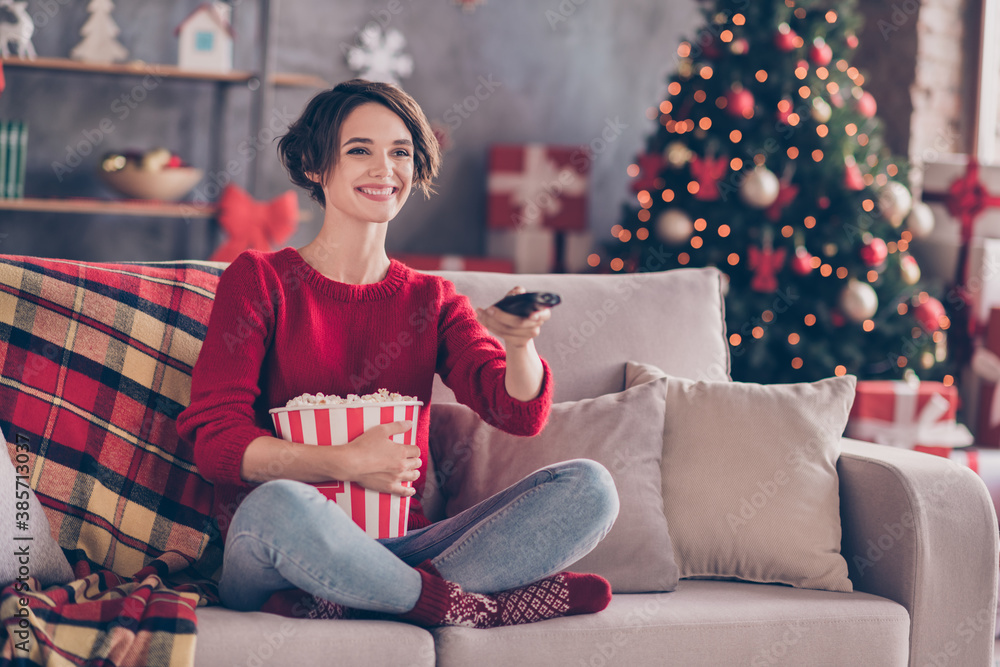 Photo of lovely young lady hold remote control embrace popcorn paper box sitting couch wear red sweater jeans socks indoors - obrazy, fototapety, plakaty 