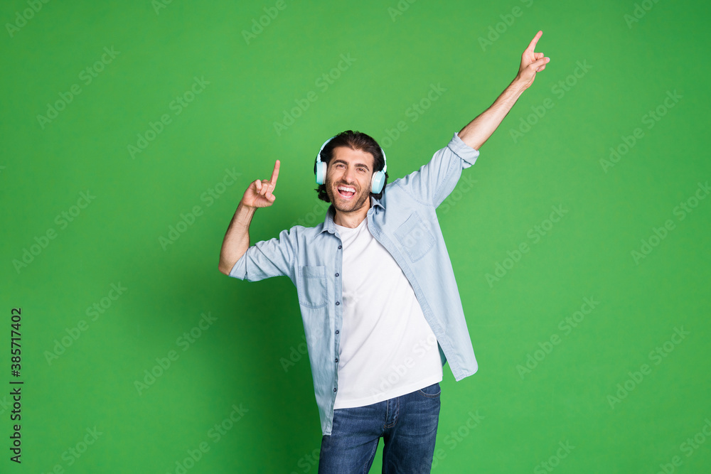Photo of stubbled young guy raise hands indicate empty space wear headphones denim shirt jeans isolated green color background Photo of stubbled young guy raise hands indicate empty space wear headphones denim shirt jeans isolated green color background