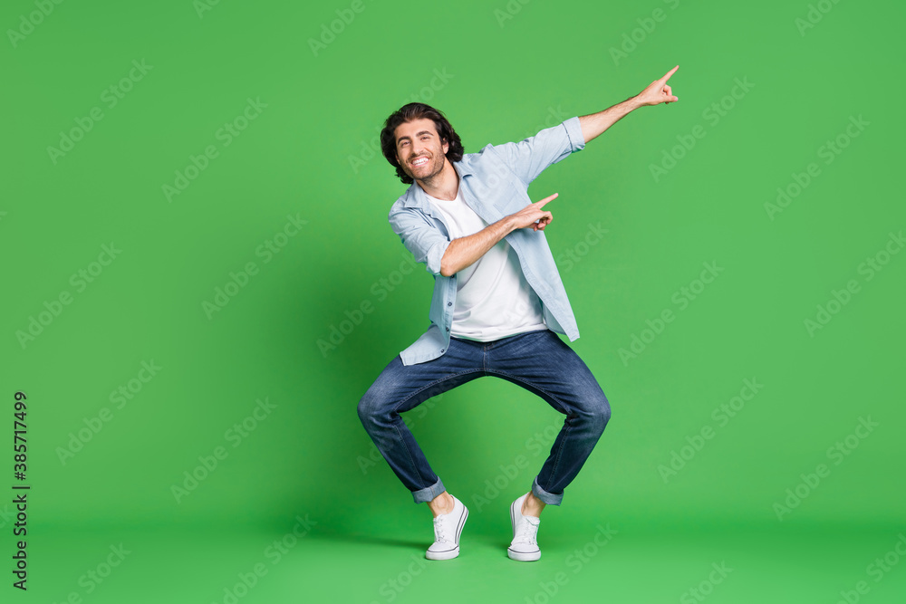 Full length photo of stubbled man funny legs direct finger empty space wear denim shirt jeans sneakers isolated green color background Full length photo of stubbled man funny legs direct finger empty space wear denim shirt jeans sneakers isolated green color background