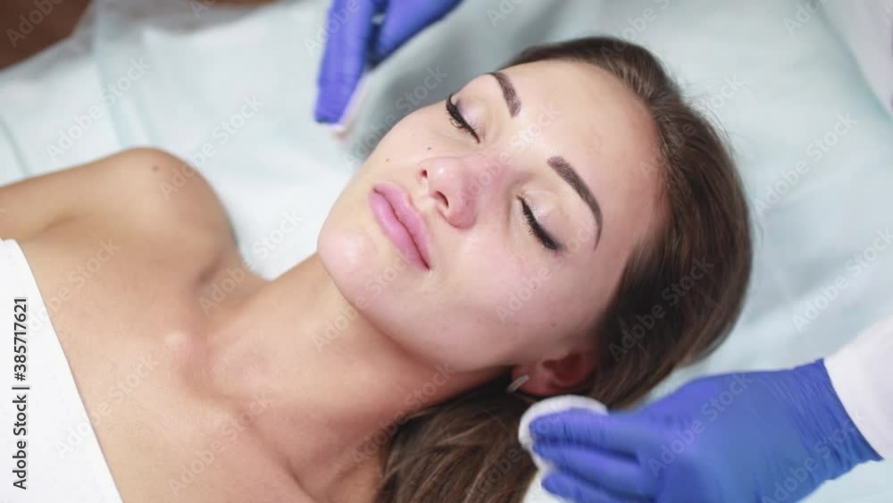 The beautician cleans the face of a young beautiful woman with cotton sponges in preparation for the subsequent procedures in the beauty salon. Cosmetology and spa.