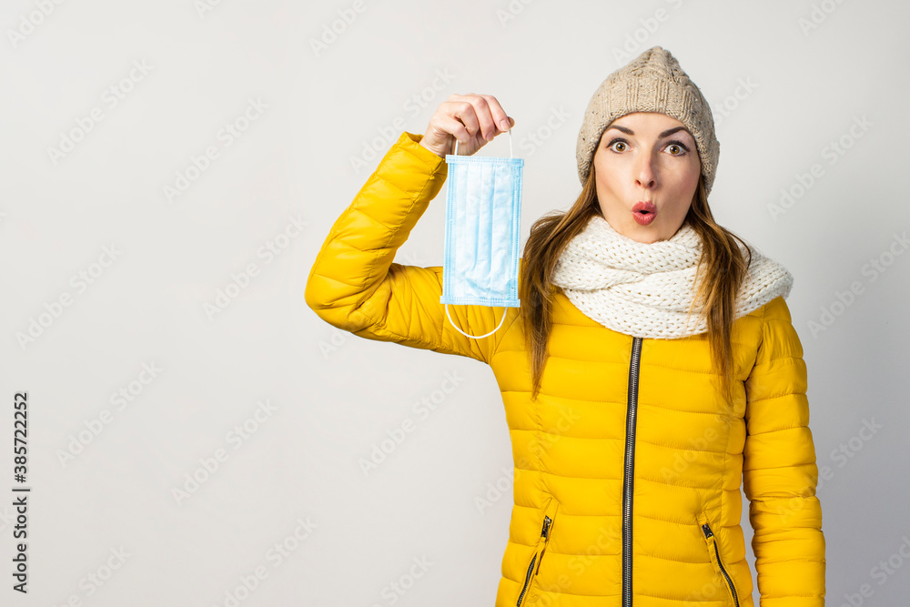 surprised young girl in a yellow jacket and hat, holds a medical mask on a light background. Concept of the common cold, virus, infectious diseases surprised young girl in a yellow jacket and hat, holds a medical mask on a light background. Concept of the common cold, virus, infectious diseases