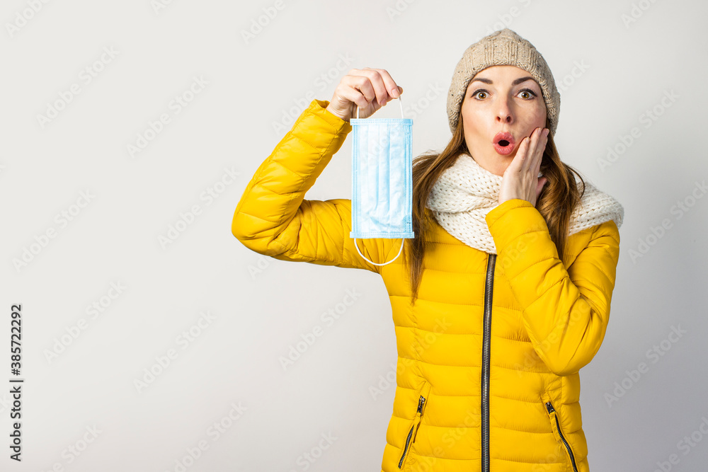 surprised young girl in a yellow jacket and hat, holds a medical mask on a light background. Сoncept of the common cold, virus, infectious diseases surprised young girl in a yellow jacket and hat, holds a medical mask on a light background. Сoncept of the common cold, virus, infectious diseases