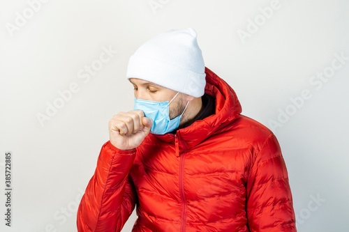 young man with a medical mask on his face coughs on a light background. Concept of the common cold, virus, infectious diseases