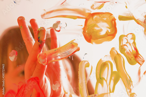 Beautiful young girl painting artwork on glass with colorful hands and finger. Happy childhood, art, painting lessons concept. Selective focus on finger. Horizontal image.