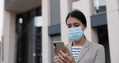 Close up portrait of beautiful Caucasian young female in medical mask walking outdoors on street and texting on cellphone. Caucasian woman browsing on smartphone and looking away. Job concept