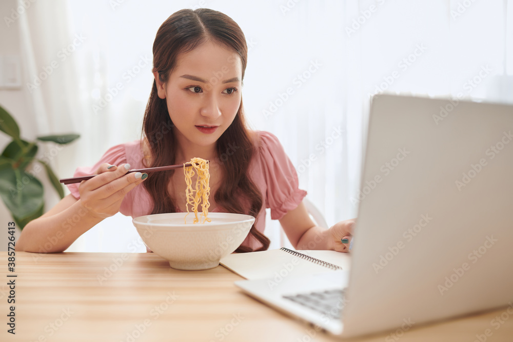 Beautiful young Asian woman using laptop and eating Asian noodles at home. Beautiful young Asian woman using laptop and eating Asian noodles at home.