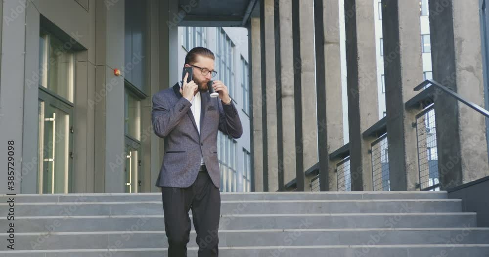 Front view of attractive successful confident adult bearded office worker in glasses and stylish suit which drinking coffee and talking on phone while leaving the office building by stairs