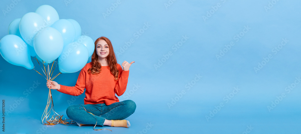 Smiling woman with long red hair in casual attire holding lots of air balloons and pointing finger aside, isolated over blue background, lady with crossed legs sitting on floor.