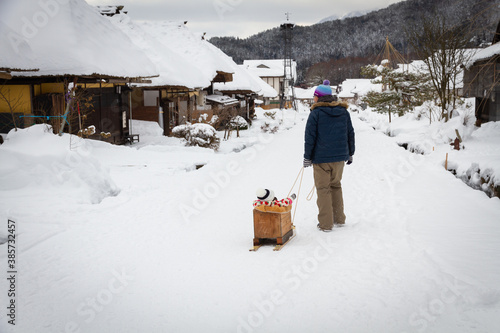 Landscape of Ouchijuku village in winter which snow covered all over the road and cottage's thatched roof and father draw a cart that a girl sit inside along the street. photo