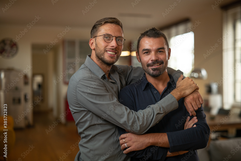 Authentic shot of young happy smiling married homosexual male gay couple is enjoying time together is hugging as sign of timeless love and smiling in camera in a kitchen at home. Authentic shot of young happy smiling married homosexual male gay couple is enjoying time together is hugging as sign of timeless love and smiling in camera in a kitchen at home.