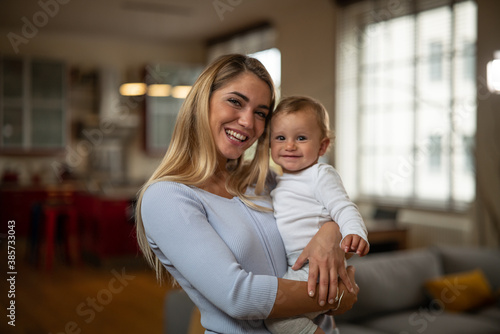 Authentic shot of an young happy mother is keeping in arms her toddler baby boy and both are smiling in camera in a living room at home.